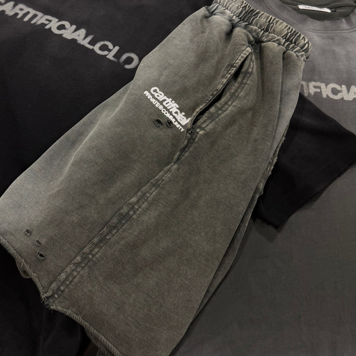 A pair of faded gray washed denim shorts with a white embroidered logo, laid flat on top of a black t-shirt. A second pair of matching gray shorts is visible underneath, along with a dark gray t-shirt in the background.