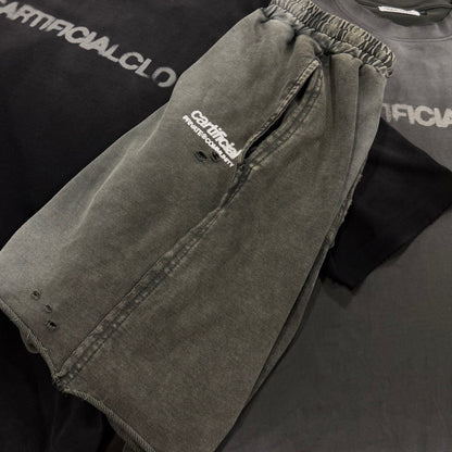 A pair of faded gray washed denim shorts with a white embroidered logo, laid flat on top of a black t-shirt. A second pair of matching gray shorts is visible underneath, along with a dark gray t-shirt in the background.