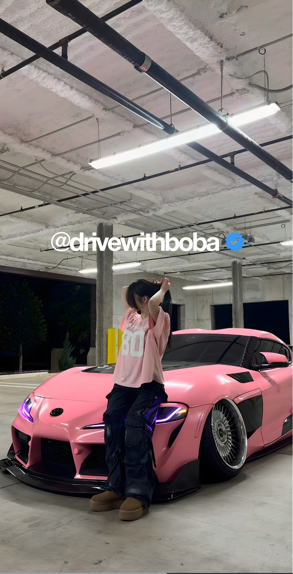 Person taking a selfie next to a pink sports car in a garage with 'drivewithboba' branding.